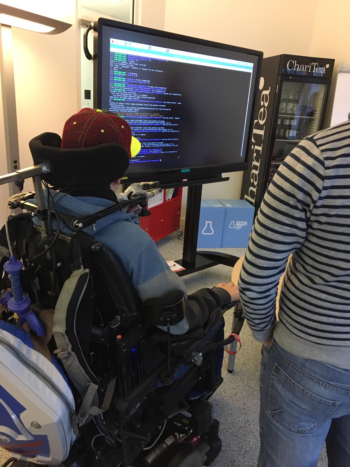 Person in a power wheelchair independently operating a large external monitor displaying code, with a collaborator standing alongside