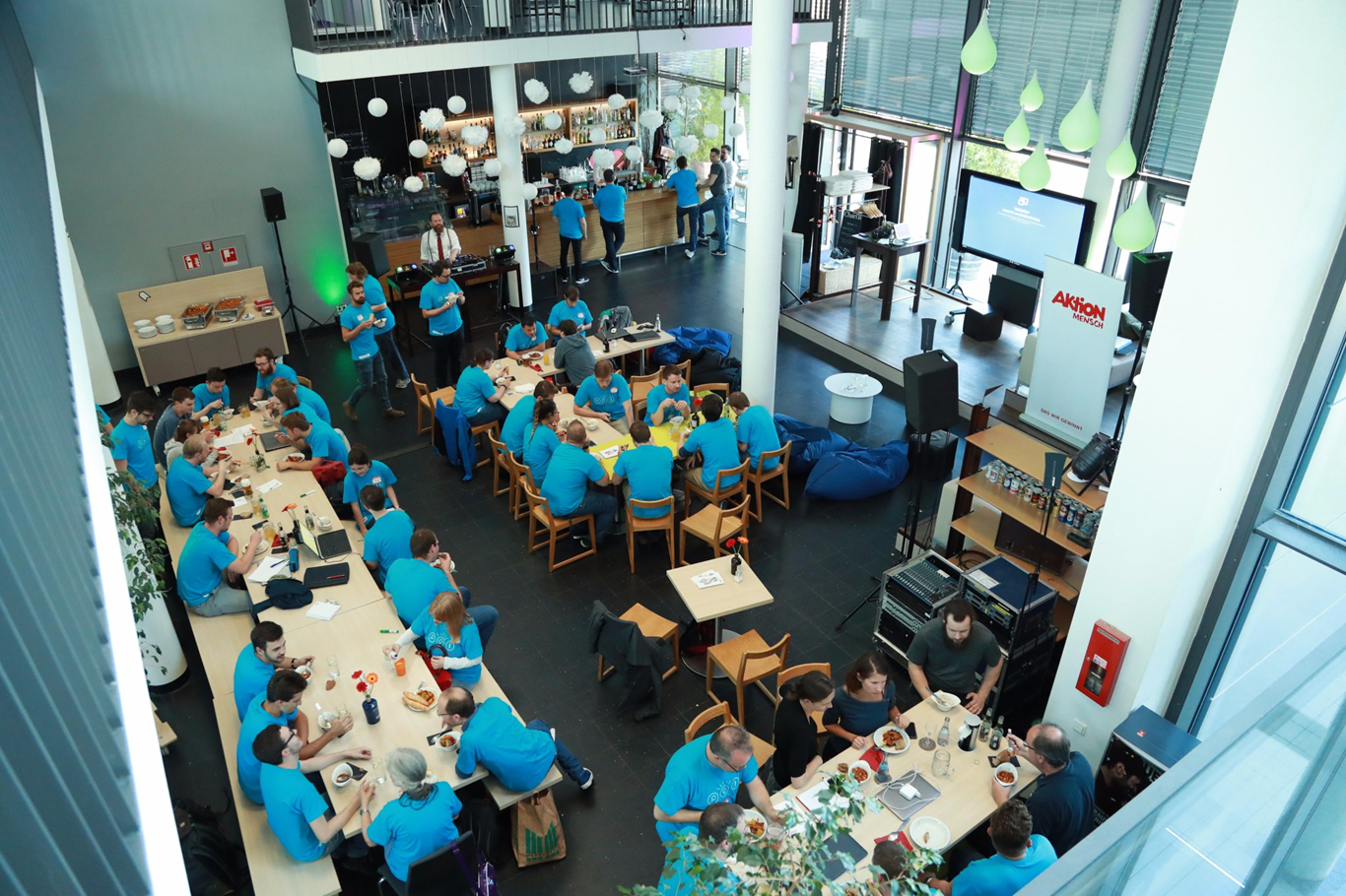 Aerial view of the BIRNE7 Neue Nähe 2.0 hackathon event organised by Aktion Mensch, with dozens of participants collaborating at tables in a large open hall